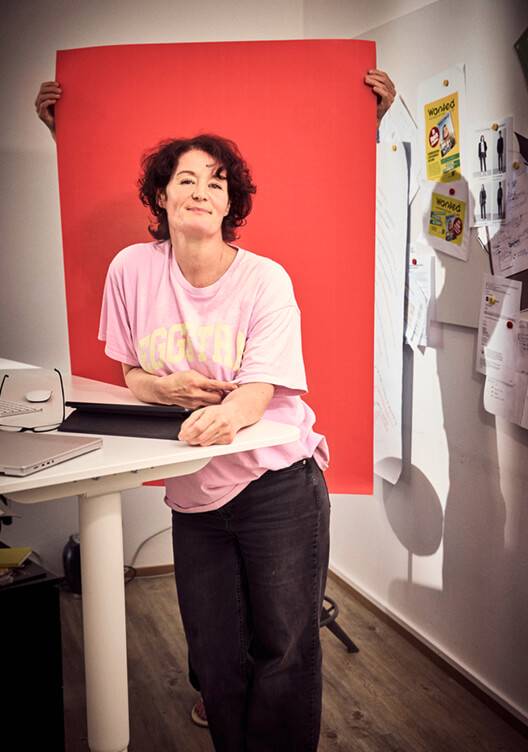 Eine Person steht in einem Raum und hält ein großes rotes Poster oder eine Leinwand. Sie lächelt und trägt ein buntes T-Shirt. Im Hintergrund sind verschiedene Gegenstände und Werkzeuge an der Wand befestigt.