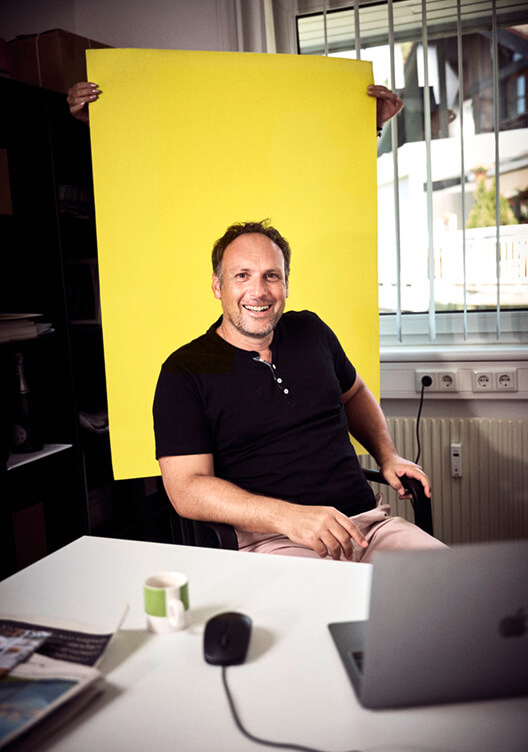 Ein Mann sitzt lächelnd an einem Schreibtisch mit einem Laptop. Im Hintergrund steht ein großer gelber Karton. Der Raum wirkt wie ein Büro oder Arbeitsbereich mit Regalen und Küchenelementen.
