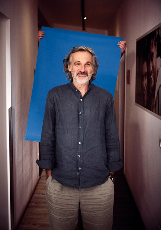 Ein Mann mit grauem Haar und grauem Hemd steht in einem Flur und hält ein großes blaues Schild. An der Wand hinter ihm hängen Bilder. Der Flur ist schmal und hat eine helle Beleuchtung.