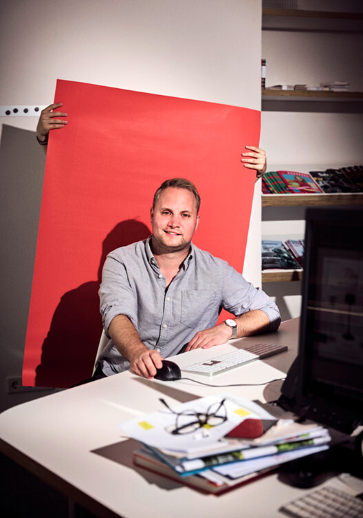 Ein Mann sitzt an einem Schreibtisch und hält ein großes rotes Blatt Papier. Er lächelt und trägt ein blaues Hemd. Auf dem Tisch befinden sich verschiedene Gegenstände wie Bücher, ein Laptop und ein Monitor. Im Hintergrund sind Regale mit Ordnern und Büchern zu sehen.