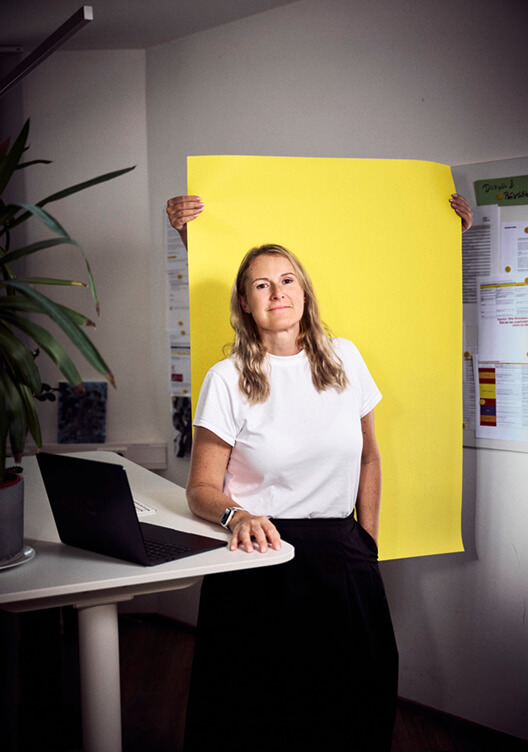 Eine Frau steht in einem Büro und hält ein großes gelbes Blatt Papier. Sie trägt ein weißes T-Shirt und eine schwarze Hose. Im Hintergrund sind ein Schreibtisch mit einem Laptop, eine Pflanze und ein Kühlschrank zu sehen.