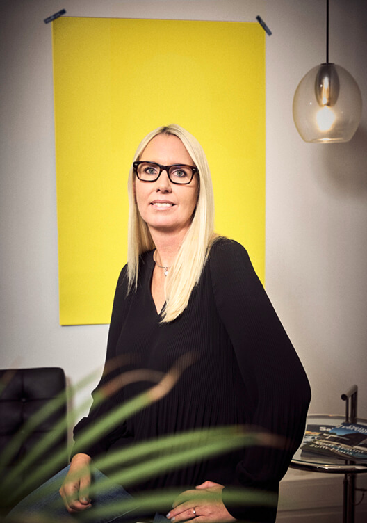 Eine Frau mit blonden Haaren und Brille sitzt in einem Raum mit einer gelben Wand und einer modernen Lampe. Sie trägt ein schwarzes Kleid und sitzt entspannt auf einem Stuhl. Im Vordergrund befindet sich eine Pflanze.