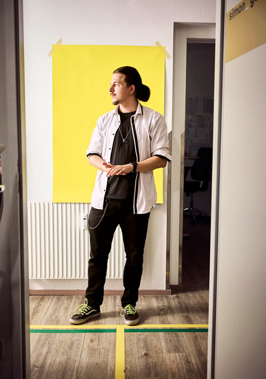 Ein Mann in weißem Hemd und schwarzem Shirt steht in einem Flur vor einem gelben Poster. Er trägt eine schwarze Hose und steht auf einem mit grünem Klebestreifen markierten Boden. Im Hintergrund ist eine Tür sichtbar.