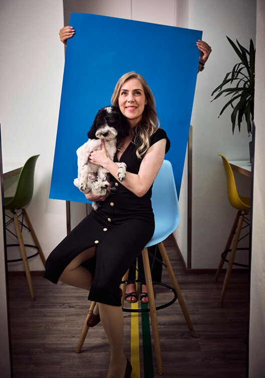 Eine Frau sitzt auf einem Stuhl und hält einen kleinen Hund. Sie lächelt und hält eine große blaue Leinwand hinter sich. Im Hintergrund sind moderne Möbel und eine Pflanze zu sehen. Die Atmosphäre wirkt freundlich und entspannt.