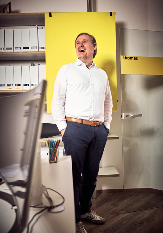 Ein Mann steht lachend in einem Büro, mit einem weißen Hemd und blauen Hosen. Im Hintergrund sind Regale mit Ordnern und ein gelbes Schild mit dem Namen "Thomas". Ein Schreibtisch mit Stuhl und Büroartikeln ist ebenfalls sichtbar.
