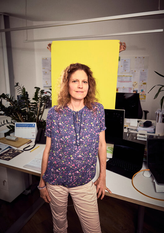 Eine Frau steht in einem Labor und trägt eine blaue Bluse mit Blumenmuster. Hinter ihr sind verschiedene Laborgeräte und ein gelbes Poster an der Wand zu sehen. Sie wirkt konzentriert und steht mit verschränkten Armen.
