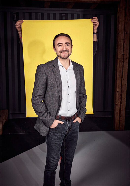 Ein Mann steht lässig mit den Händen in den Taschen vor einem gelben Hintergrund. Er trägt einen Tweed-Blazer, ein weißes Hemd und eine Jeans. Das Bild wirkt professionell und zeigt ihn in einem gut beleuchteten Raum.